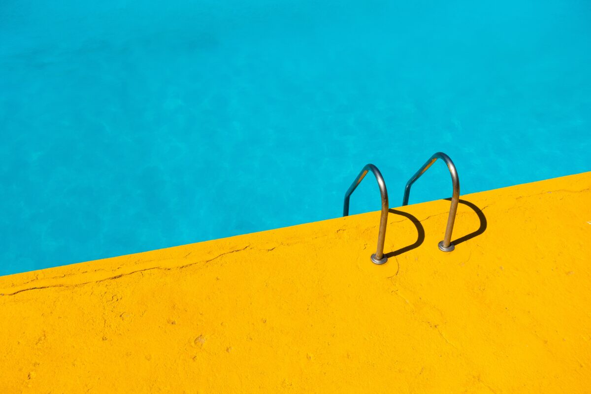 Photograph of the top of a metal ladder at the edge of a pool. The edge of the pool is yellow, contrasts with the blue of the water.