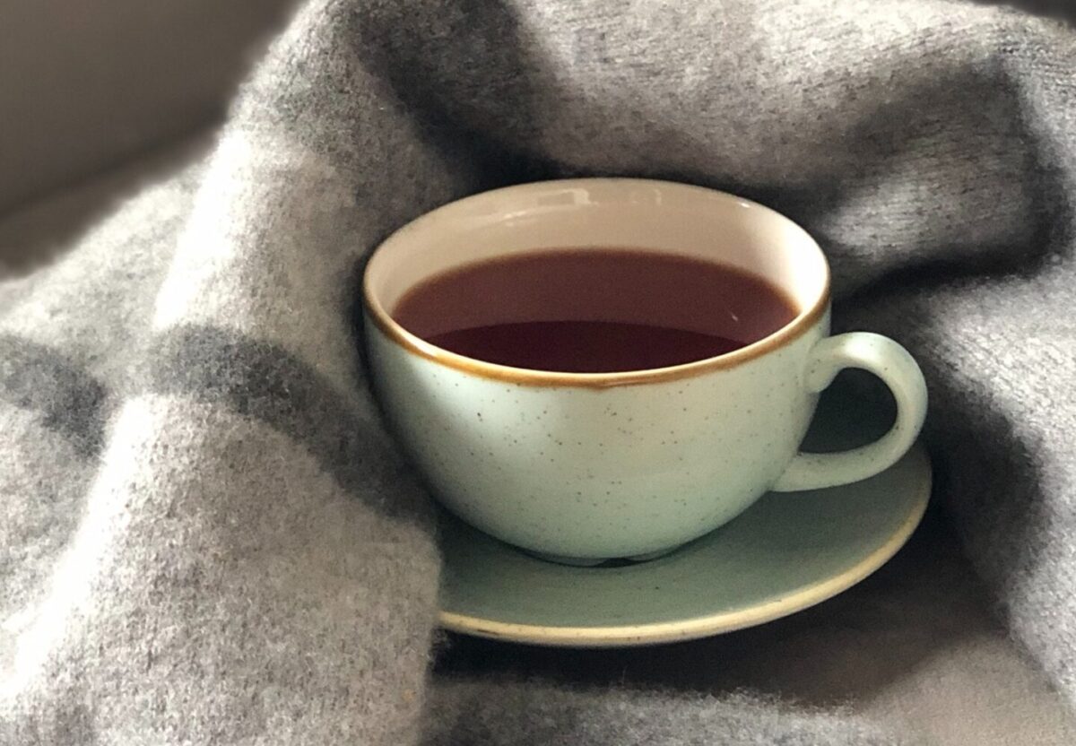 Photograph of a cup of coffee, or tea, with a grey wool blanket in the background.